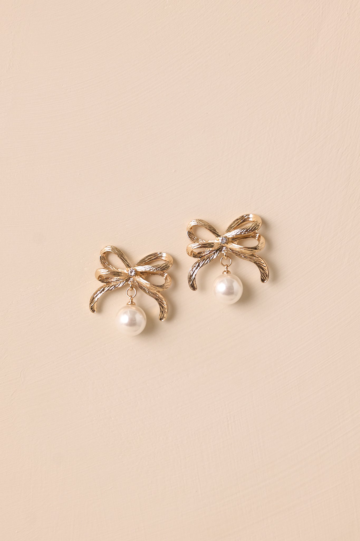 A pair of gold and ivory bow earrings displayed against a beige background, featuring a gold bow with rhinestone detailing and a large ivory faux pearl hanging below.