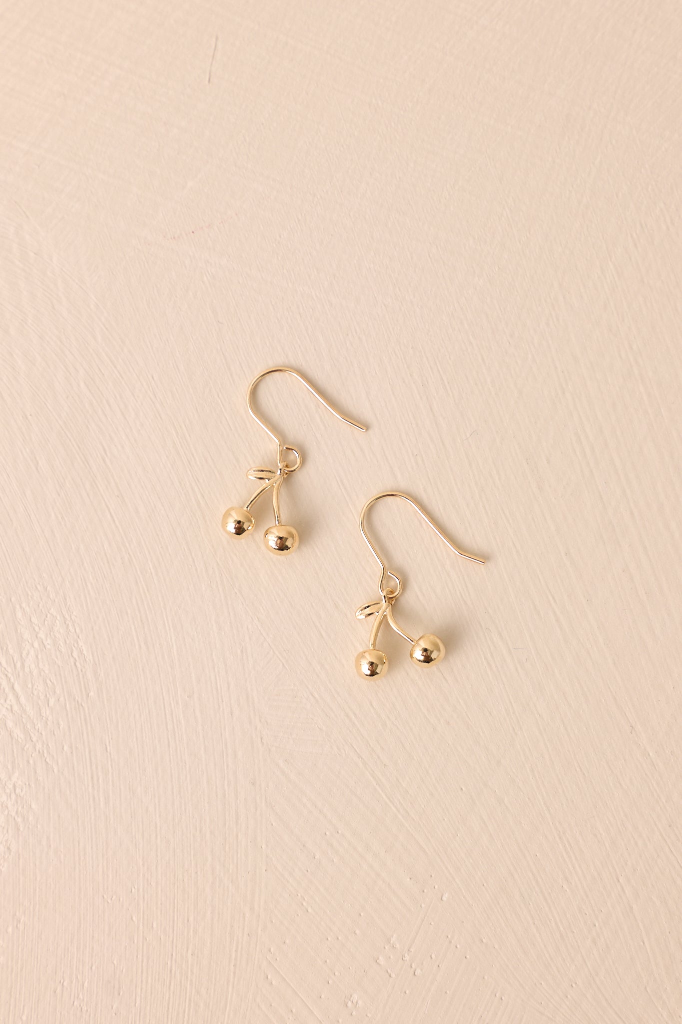 These gold earrings showcase cherry-shaped charms dangling from fish hook wires. The polished gold finish enhances their timeless look, with clear silicone backings for added stability.