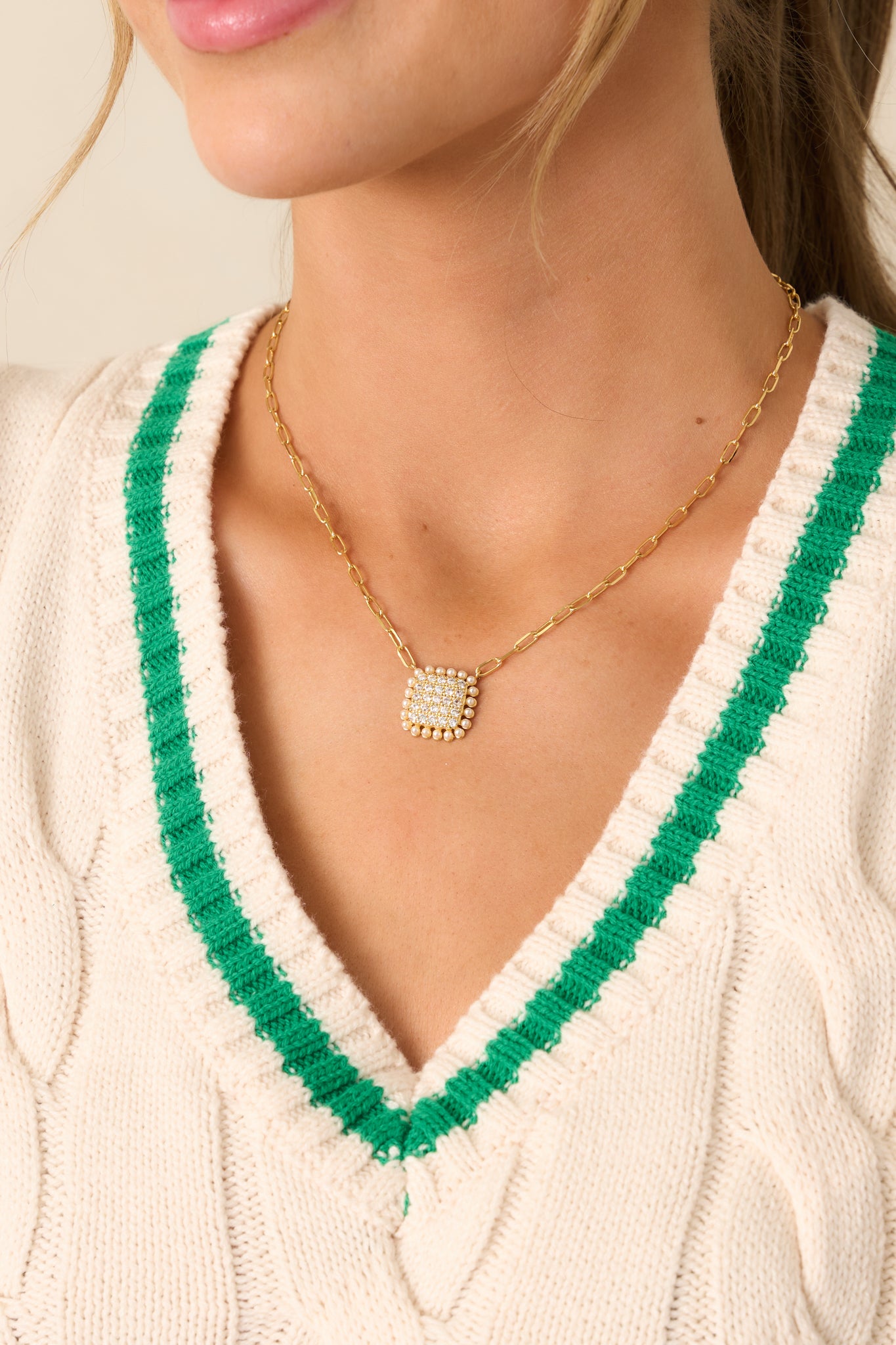 This gold necklace features a sleek rectangular chain and a statement square pendant, embellished with rhinestones and surrounded by an elegant ivory faux pearl border.