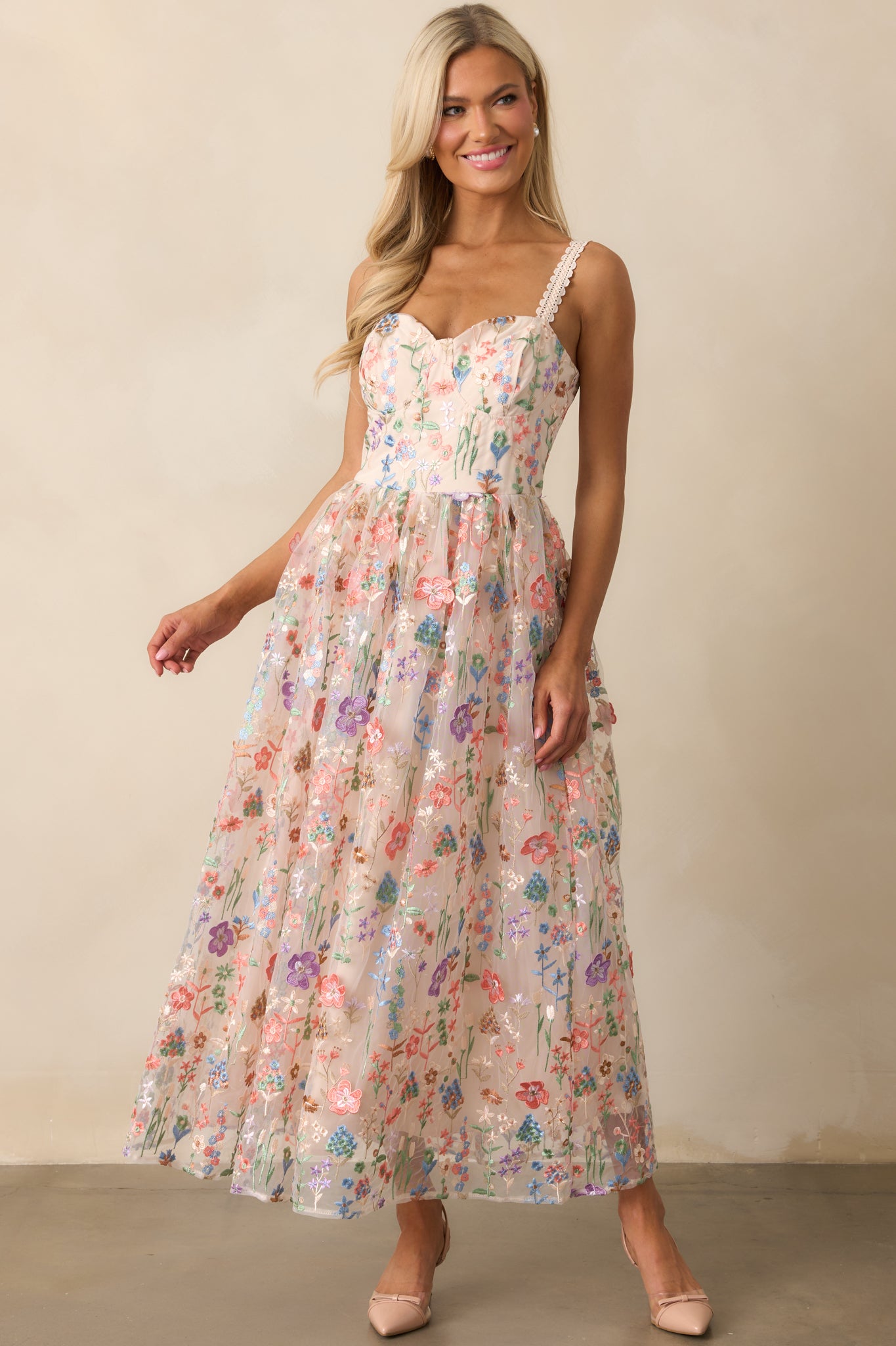 A rose pink floral dress with a rounded balconette cup neckline, fitted bodice, and a relaxed skirt featuring a sheer floral embroidered overlay. The dress is shown from the front, highlighting its elegant design.