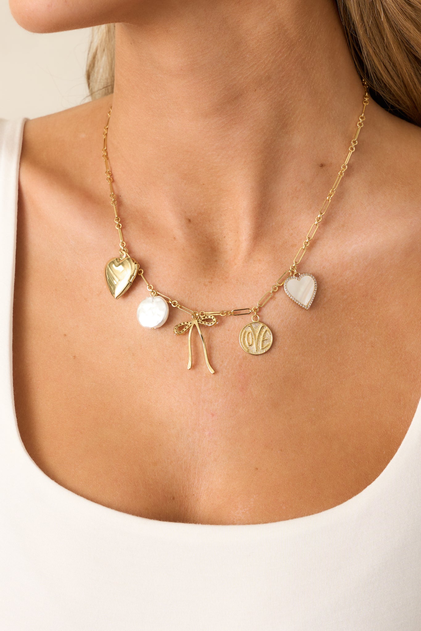 A gold heart charm necklace with a thin rectangular chain, featuring a mix of gold and ivory charms, including a gold heart locket and a pearlescent circular charm.