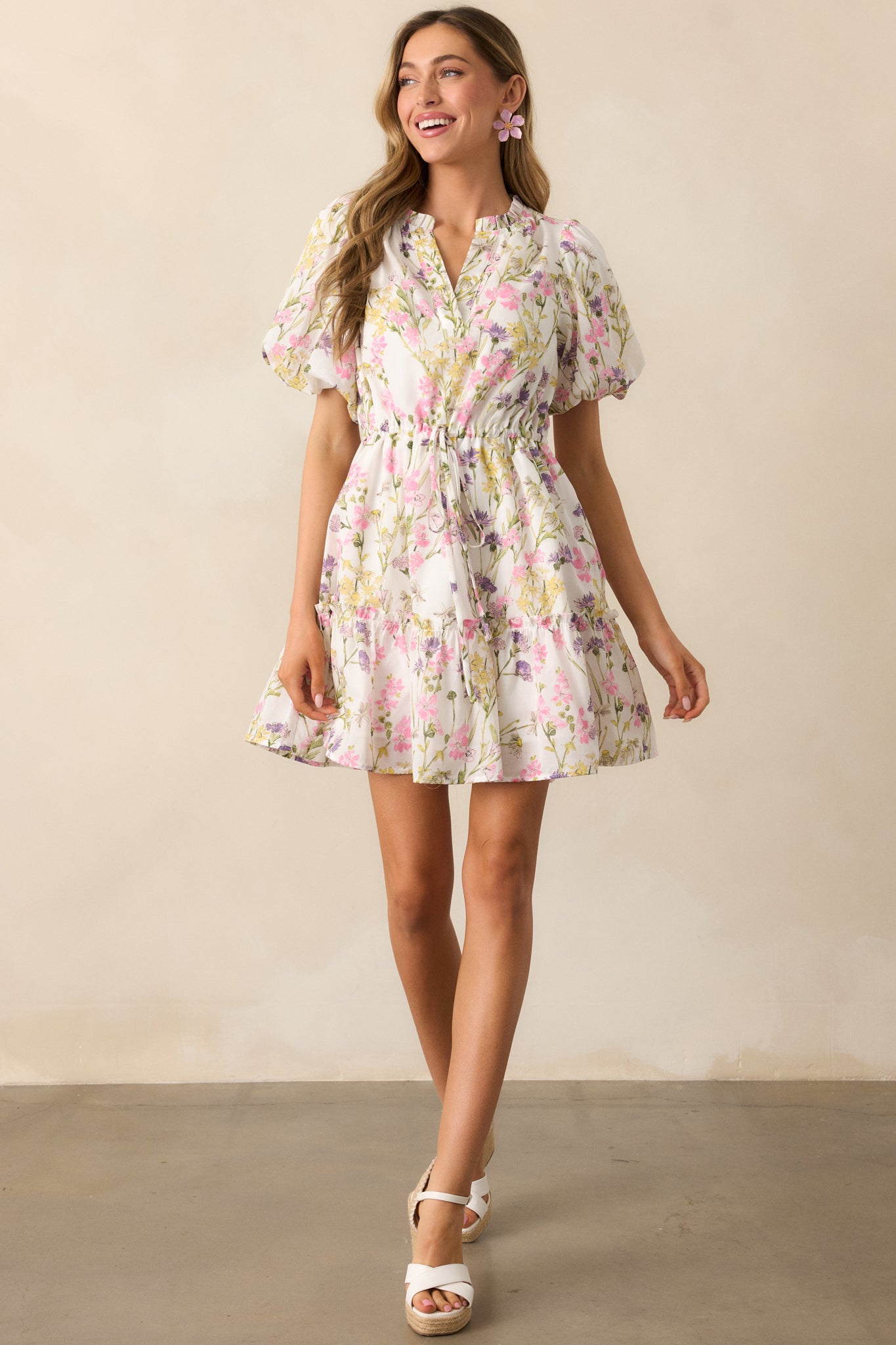 This ivory mini dress features a pink multicolor floral print, a notch v-neckline, and short puff sleeves with elastic cuffs. The adjustable self-tie drawstring waist and ruffle details add a feminine touch, while functional pockets complete the look.