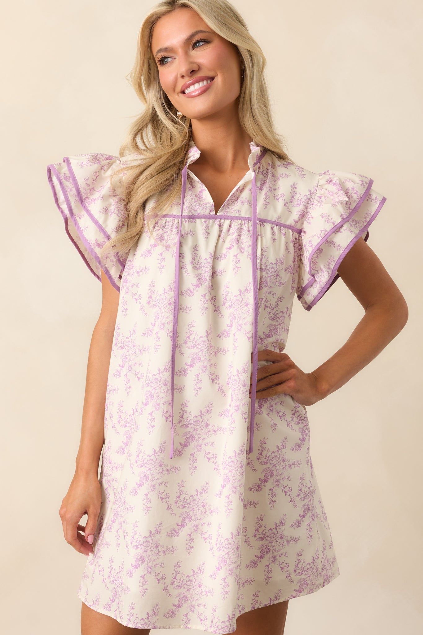 A charming mini dress with a delicate lavender toile print, flutter sleeves, and a ruffle collar with an optional self-tie for a touch of elegance.


