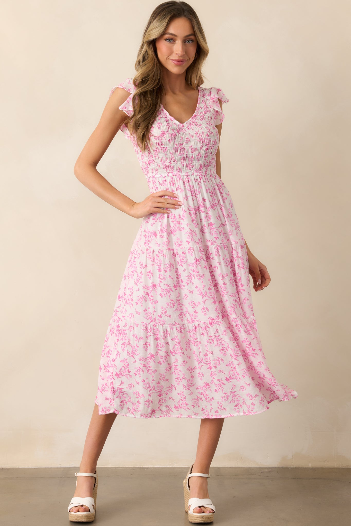 This pink floral dress features a flattering v-neckline, a fully smocked bust, and flutter sleeves that add a feminine touch. The tiered skirt flows elegantly, with an ivory lining providing a soft contrast.
