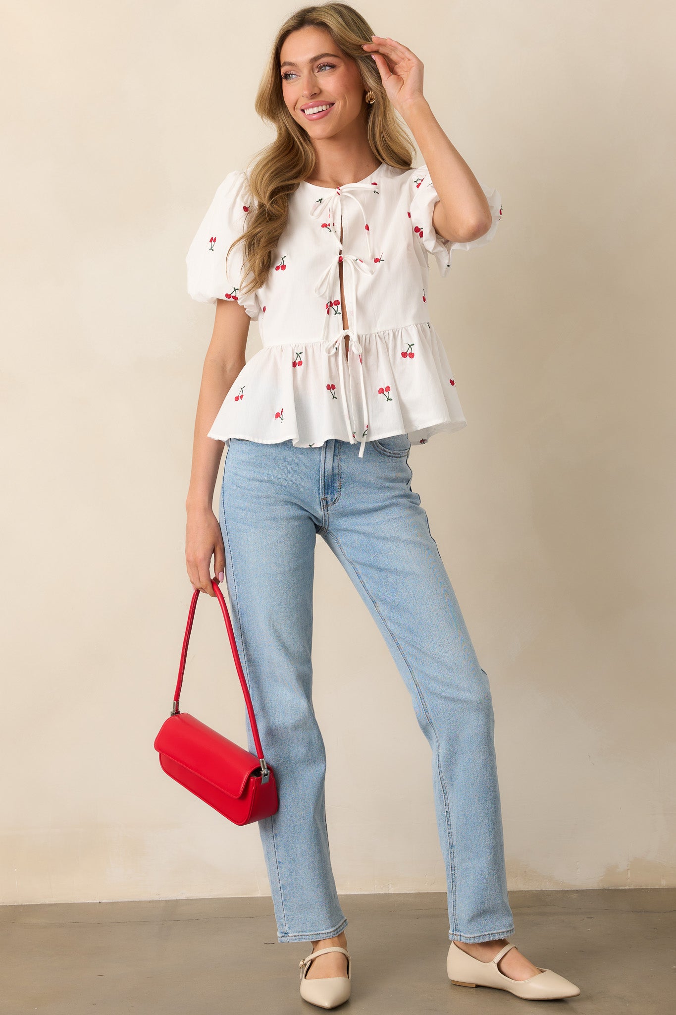 A white top adorned with embroidered cherry icons, featuring three adjustable self-tie bows down the front, short puff sleeves with elastic cuffs, and a playful peplum silhouette.

